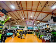 Innenansicht einer großen Autowerkstatt mit hoher Decke und sichtbaren Trägern, orangefarbenem Boden und breiten Fenstern entlang der Rückwand. Mehrere Hebebühnen mit Fahrzeugen sowie Werkbänke, Regale und Werkzeugwagen füllen die Halle, beleuchtet von großen, rasterförmigen Deckenleuchten.