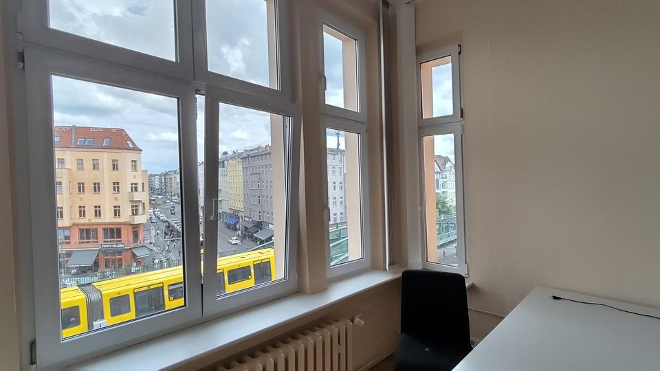 Heller Büroraum mit großen, weiß gerahmten Fenstern und Blick auf eine städtische Kreuzung mit mehrstöckigen Gebäuden und einer gelben Straßenbahn. Im Inneren sind eine weiße Tischplatte, ein schwarzer Stuhl und ein Heizkörper unter der Fensterbank zu sehen.