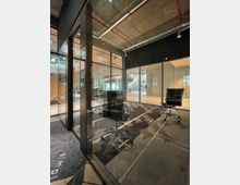 Modernes Büro mit verglasten Besprechungsräumen, zwei schwarzen Bürostühlen und geometrischem Teppichmuster auf dem Boden. Im Hintergrund offene Sicht auf eine industrielle Decke mit freiliegenden Rohren und Treppen in einem offenen Raum.