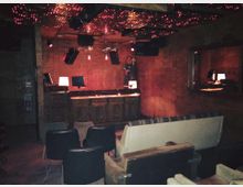 Ein kleiner Raum mit gedämpfter Beleuchtung, ausgestattet mit verschiedenartigen Sesseln und Sofas. Im Hintergrund befindet sich eine hölzerne Bar oder DJ-Pult mit computergesteuerten Elementen, roten Beleuchtungseffekten und einem funkelnden Sternenhimmel an der Decke.