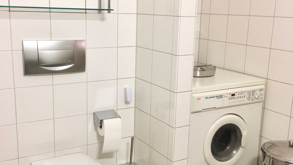 Kleines, weiß gefliestes Badezimmer mit wandhängendem WC, Spülkasten-Bedienfeld, Glasablagen und Toilettenpapierhalter. Rechts steht in einer Nische eine Frontlader-Waschmaschine neben einem Metall-Wäschekorb auf hellen Bodenfliesen.