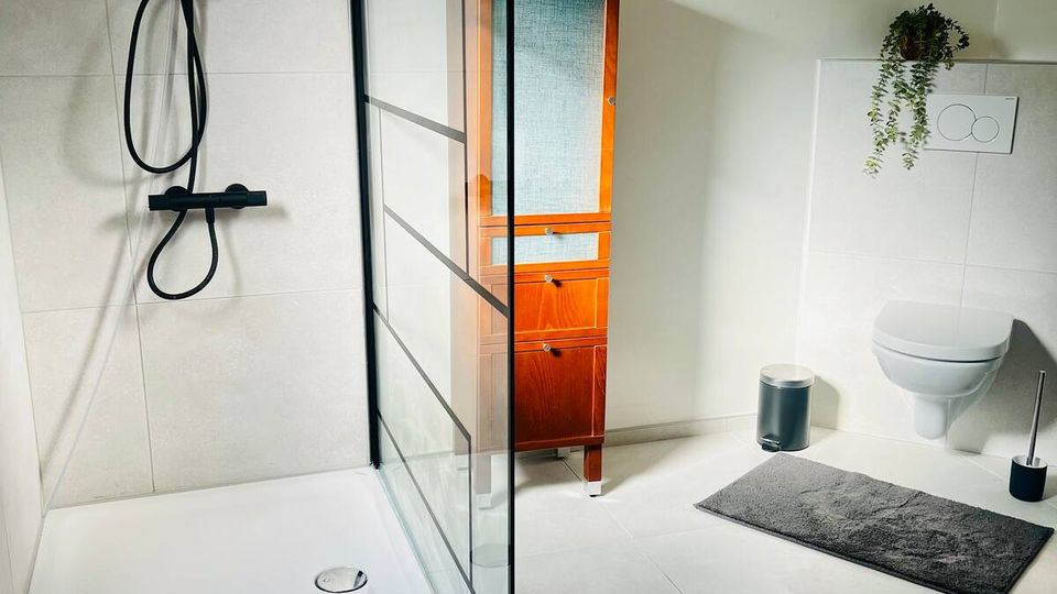 Modernes Badezimmer mit bodengleicher Dusche, schwarzer Regendusche und Glas-Duschabtrennung mit schwarzen Rahmen. Rechts sind ein wandhängendes WC mit Betätigungsplatte, ein Holzschrank sowie helle Fliesen und graue Badvorleger zu sehen.