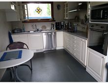 Helle Küche mit weißen Schränken, grauer Arbeitsplatte und modernen Edelstahlgeräten wie einem Backofen, einer Mikrowelle und einer Spülmaschine. Ein rundes weißes Tischchen mit einem Stuhl steht vor dem Fenster, das mit einem bunten Glasornament verziert ist.