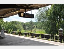 Ein Bahnsteig mit überdachter Wartehalle in einer ländlichen Umgebung, umgeben von Bäumen und einer weitläufigen Grünfläche. Eine Uhr und eine Anzeigetafel hängen unter dem Holzdach, während das Bahnsteigende von einer niedrigen Ziegelmauer gesäumt ist.