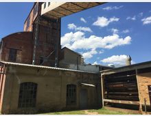 Industriegebäude mit Backsteinfassade und Metallkonstruktionen, verbunden durch einen erhöhten Steg. Teile des Gebäudes zeigen verfallene Fenster, Wellblechdächer und offene Stahlstrukturen, umgeben von einer Wiese unter blauem Himmel mit vereinzelten Wolken.