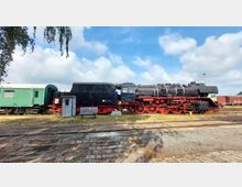 Historische Dampflokomotive mit schwarzem Kessel und roten Rädern, abgestellt auf Gleisen neben einem grünen Waggon. Im Vordergrund stehen eine Sitzbank und ein kleines Metallhäuschen, im Hintergrund sind weitere Waggons und der Himmel mit einigen Wolken zu sehen.