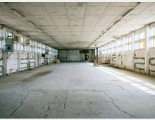 Weitläufiger, leerer Industrie- oder Lagerraum mit einer Betonbodenfläche, sichtbaren Rissen und hohen Wänden mit gleichmäßig angeordneten Fenstern, die Tageslicht hereinlassen. Entlang der Wände verlaufen Rohre und Leitungen, die den funktionalen Charakter des Raums hervorheben.