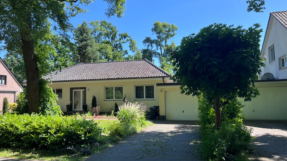 Einfamilienhaus mit flachem Walmdach und heller Fassade in einer grünen Wohnstraße, davor Gartenbeete und ein gepflasterter Zufahrtsweg. Rechts schließt eine Garage an, umgeben von großen Bäumen und dichtem Strauchwerk.