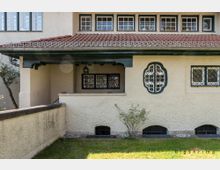 Ein kleiner Bereich einer historischen Villa mit verputzten Wänden, einem roten Ziegeldach und verzierten Fenstern mit Gitterelementen. Im Vordergrund ein Grünstreifen, kleine Sträucher und eine niedrige Mauer.