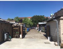 Hinterhof mit gepflastertem Weg, flankiert von einfachen Schuppen aus Holz und Ziegeln. Im Hintergrund sind Bäume sowie Dächer von Wohnhäusern und ein Kirchturm zu sehen.