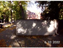 Straßenszene mit herbstlichem Laub auf Gehwegen; im Vordergrund eine weiße Mauer, dahinter Bäume und ein Gebäude mit roter Klinkerfassade und großen Fenstern.