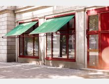 Fassade eines Gebäudes mit großen Fenstern und roten Rahmen, teilweise mit grünen Markisen bedeckt; rechts befindet sich eine Glastür mit einer Aufschrift und einem goldenen Türgriff. Der Boden davor ist mit Kopfsteinpflaster belegt, Bäume und parkende Autos sind im Hintergrund erkennbar.