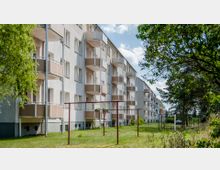 Mehrstöckiges Wohngebäude im Plattenbaustil mit beige-braunen Balkonverkleidungen, umgeben von einer Grünfläche mit Bäumen und Wäscheleinen im Vordergrund. Der Himmel im Hintergrund ist leicht bewölkt.