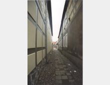 Schmale, gepflasterte Gasse mit Fachwerkhäusern auf beiden Seiten; die Häuser haben helle Wände und sichtbare Holzbalkenstrukturen. Am Ende der Gasse sind weitere Gebäude und rote Ziegeldächer erkennbar.