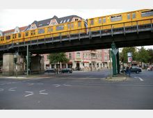 Straßenszene mit einer erhöhten Bahntrasse, auf der eine gelbe U-Bahn fährt. Darunter eine Kreuzung mit Ampeln, Fußgängern, Autos und einem Gebäude mit mehreren Geschäften, darunter eine Sparkassenfiliale.