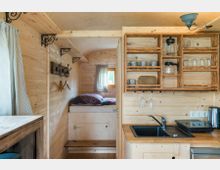 Kompakter Innenraum eines Tiny Houses mit einer Küchenzeile aus hellem Holz, offenen Regalen mit Geschirr und einer schwarzen Spüle. Im Hintergrund ist ein Schlafbereich mit einem hohen Bett und einem kleinen Fenster zu sehen.