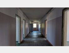 Ein leerer, schlichter Flur eines Gebäudes mit heller Wandfarbe und Fliesenboden. Auf beiden Seiten befinden sich offene Türrahmen, und am Ende des Flurs ist ein Fenster sichtbar, durch das Tageslicht einfällt.