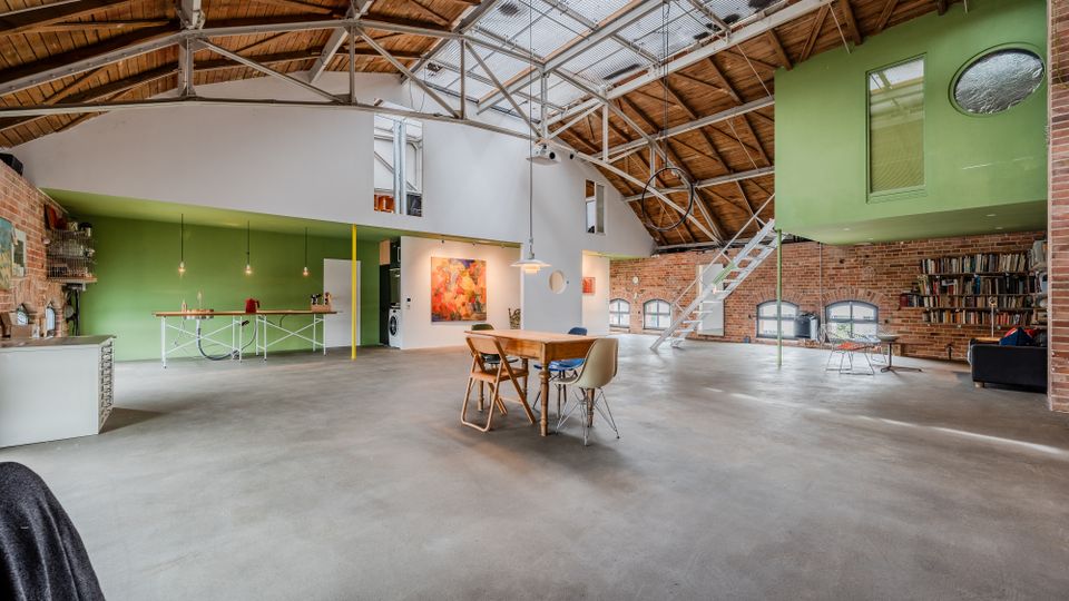 Großer, offener Loft‑Innenraum in einer umgebauten Industriehalle mit Sichtziegelwänden, Betonboden und hoher Holzdachkonstruktion mit Stahlträgern und Oberlichtern. In der Mitte stehen Tisch und Stühle, rechts führt eine Metalltreppe zu einem grün gestrichenen Zwischengeschoss, daneben Regale und Sitzbereich.