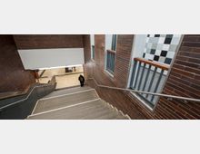 Treppe in einem öffentlichen Innenraum mit Wänden aus roten Backsteinen, Metallgeländern und kleinen Fenstern mit schwarz-weiß-karierten Fliesen. Unten erkennt man einen Flur mit hellen Fliesen und eine Person mit Rucksack.