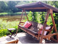 Holz-Schaukelbank mit Überdachung und Kissen, umgeben von grünen Pflanzen, auf einer Steinterrasse neben einem kleinen Teich in einer natürlichen Umgebung. Ein kleiner, klappbarer Holztisch steht daneben.