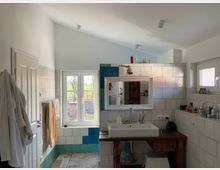 Helles Badezimmer mit weiß gefliesten Wänden, einem rechteckigen Waschbecken auf einem rustikalen Holztisch und einem großen Fenster mit Blick ins Freie. Auf der linken Seite hängen Handtücher und Bademäntel an einer Tür, daneben sind blaue und weiße Fliesen im unteren Bereich der Wand angebracht.