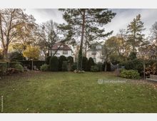 Weitläufiger Garten mit Rasenfläche und mehreren Bäumen, umrahmt von Hecken und Sträuchern. Im Hintergrund stehen zwei helle Häuser mit Satteldächern, umgeben von einer natürlichen, herbstlichen Vegetation.
