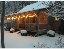 Holzhaus mit überdachter Veranda, dekoriert mit leuchtenden Lichterketten, umgeben von einem verschneiten Garten mit vereisten Büschen und Bäumen. Im Hintergrund steht ein Wald, ebenfalls mit Schnee bedeckt.