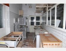 Verlassene Küche mit weißen Fliesenwänden, Metallspüle, Arbeitsflächen und leeren Regalen. Der Raum wirkt ungepflegt, mit abgenutztem Boden und ungenutzten Geräten.