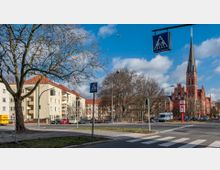 Straßenszene mit einer Kreuzung im Vordergrund, einer Fußgängerampel und einem Zebrastreifen. Im Hintergrund ein rotes, neugotisches Kirchengebäude mit spitzem Turm, umgeben von Bäumen, sowie Wohngebäude mit hellen Fassaden und roten Dächern. Ein Flugzeug ist am blauen Himmel sichtbar.