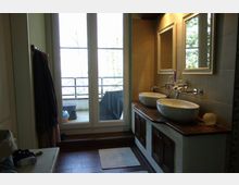 Badezimmer mit zwei runden Waschbecken auf einer Holzplatte, darüber zwei Spiegel mit Rahmen. Im Hintergrund eine große Glastür, die auf einen Balkon mit Sitzmöbeln und Holzgeländer führt.