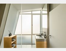 Minimalistisch eingerichtetes Badezimmer mit großem, mattiertem Fenster, einem rechteckigen Waschbecken mit Holzunterschrank und einem schmalen Regal mit Handtüchern und einem Duftspender. Die Wände und Bodenfliesen sind in hellen, neutralen Farben gehalten.