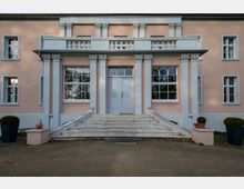 Fassade eines historischen Gebäudes mit pastellrosa Putz und weißen Details, symmetrisch angeordneten Säulen mit dekorativen Kapitellen, großen Sprossenfenstern und einer breiten Treppe, die zu einer zentralen Eingangstür führt. Links und rechts befinden sich Pflanzen in großen Töpfen.