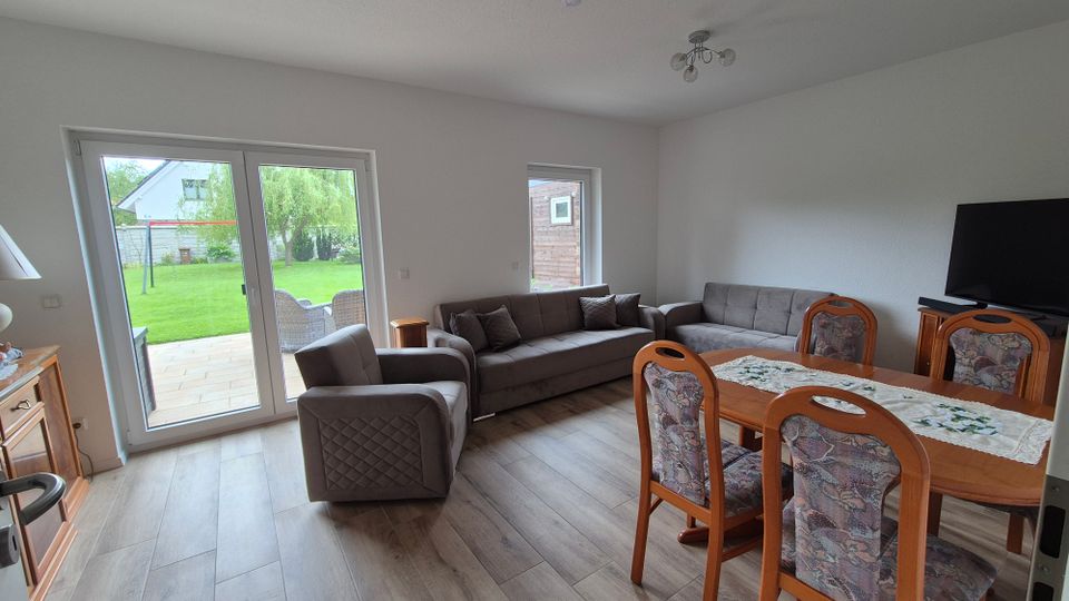 Helles Wohnzimmer mit Essbereich, grauem Bodenbelag und Sitzgruppe aus Sofa und Sessel; nebenan steht ein Esstisch mit gepolsterten Stühlen und ein Fernseher auf einem Lowboard. Große Terrassentüren führen auf eine gepflasterte Terrasse und in einen grünen Garten.