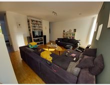 Wohnzimmer mit Holzboden, zwei dunklen Sofas, einem Couchtisch und einem großen Regal mit Fernseher, Lautsprechern und Büchern an der Wand. Im Hintergrund befindet sich eine weiße Wand mit weiteren Bücherregalen und dekorativen Elementen.