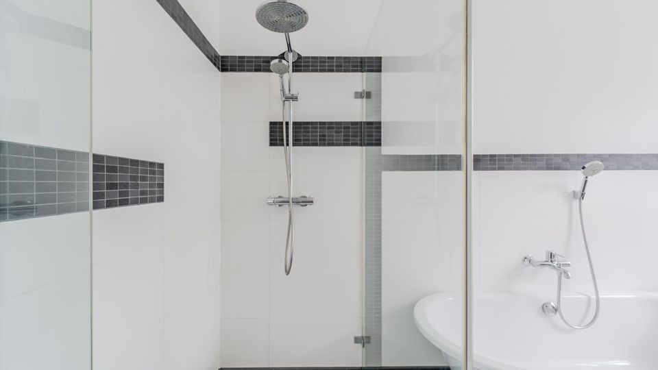 Modernes Badezimmer mit bodengleicher Dusche hinter Glaswand, weiß gefliest und mit dunklem Fliesenband akzentuiert. Rechts ist eine freistehende Eckbadewanne mit Handbrause und Armatur zu sehen.