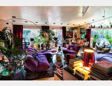 Wohnzimmer mit farbenfroher Einrichtung, lila Polstermöbeln, zahlreichen Zimmerpflanzen und dekorativen Elementen wie Wandbildern und Büchern. Große Fenster und Schiebetüren lassen viel Tageslicht herein, ergänzt durch moderne Beleuchtung und einen hängenden Acrylsessel.