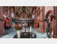 Gotisches Kircheninnere aus Backstein mit hohen Gewölbedecken, dekorativen Bögen und einer Zwischen-Empore. Im Vordergrund ein Tisch mit einem Blumenstrauß, einem Holzkreuz und einer Kerze; rechts eine moderne Skulptur aus Metall.