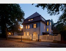 Zweistöckiges Wohnhaus mit weiß verputzter Fassade und rotem Satteldach, umgeben von einem weißen Lattenzaun. Die Fenster sind von innen beleuchtet, und das Gebäude steht an einer Kopfsteinpflasterstraße, flankiert von Bäumen und einem Laternenpfahl.