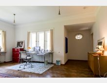 Helles Arbeitszimmer mit einem Schreibtisch vor einem Fenster, ausgestattet mit einem Computer und Lampe. Der Raum hat einen Parkettboden, einen roten Aktenschrank, einen teppichbedeckten Bereich und führt zu einem Flur mit einem kleinen ovalen Fenster.