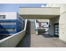 Außenbereich einer modernen urbanen Struktur mit breiter Treppe und grauen Betonwänden, teils mit Graffiti bedeckt. Links ist ein blau-weißes Bürogebäude, im Hintergrund ein rotes Firmenlogo sowie parkende Autos sichtbar.