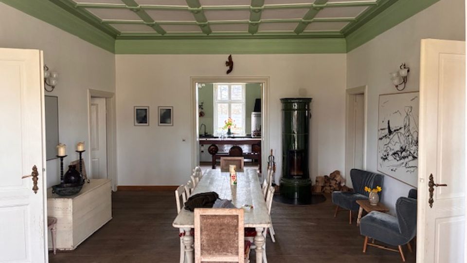 Großer, heller Essraum mit langem Esstisch und Stühlen auf Holzboden sowie grün gerahmter Kassettendecke. An den Wänden hängen Bilder; rechts stehen Sessel neben einem grünen Kachelofen und im Hintergrund ist eine Durchgangsöffnung zu einem weiteren Raum mit Fenster zu sehen.