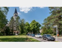 Eine Straße in einer grünen Wohngegend mit parkenden Autos, flankiert von Bäumen und einem Rasen. Im Hintergrund steht eine helle Kirche mit einem spitzen Turm und dunkelblauem Dach.