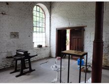 Innenraum eines alten Gebäudes mit weiß gestrichenen Ziegelwänden, einem hohen Fenster mit Metallstreben und einer Holztür im Hintergrund. Im Raum befinden sich eine traditionelle Druckpresse, ein Tisch mit kleinen Figuren, sowie weitere verstreute Gegenstände auf einem Betonboden.