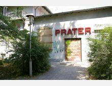 Fassade eines Gebäudes mit der roten Leuchtschrift „PRATER“ über einem Eingang, umgeben von Büschen und Bäumen. Die Wand ist teils mit Graffiti bedeckt, und neben der Tür befindet sich ein abgedecktes Fenster mit Metallgittern.