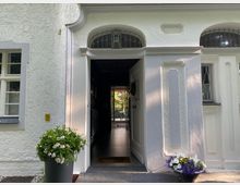 Weiße Hausfassade mit strukturiertem Putz und grauen Akzenten um die Fenster. Eine geöffnete Eingangstür führt in einen Flur mit Blick auf einen Garten im Hintergrund; Blumen in Töpfen stehen neben dem Eingang.
