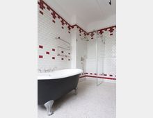 Modernes Badezimmer mit weiß gefliesten Wänden, akzentuiert durch rote Kacheln. Freistehende schwarze Badewanne mit silbernen Füßen, daneben eine Duschkabine mit Glaswand; Boden mit hellem Terrazzo-Muster.