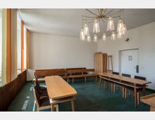 Ein einfacher Besprechungsraum mit Holztischen und -stühlen auf einem grünen Boden. Die Wände sind weiß, es gibt mehrere Holzbänke an der hinteren Wand, orange- und gelbfarbene Vorhänge an den Fenstern sowie eine große Hängelampe mit Glaslampenschirmen an der Decke.