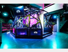 Futuristisch gestaltete Gaming-Event-Location mit einer gläsernen, beleuchteten Kabine in geometrischem Design, umgeben von farbigen Lichtakzenten. Im Hintergrund sind Zuschauertribünen und moderne Beleuchtungssysteme erkennbar.