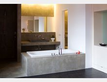 Modernes Badezimmer mit freistehender Badewanne, grauen Fliesen und einem Wandwaschbecken mit großem Spiegel. Im Hintergrund ist eine offene Dusche mit dunklen Fliesen zu sehen.