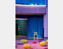 Ein moderner Innenraum mit einem blauen Vorhang im Hintergrund, vor dem ein einzelner Holzstuhl steht. Im Vordergrund liegt ein rosa Teppich, auf dem runde, bunte Sitzkissen in Gelb, Rot und Grau verteilt sind.